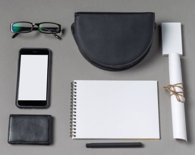 Pürüzsüz siyah bir kese, not defteri, akıllı telefon, cüzdan, gözlük ve katlanmış kağıt içeren çağdaş bir çalışma alanı. Hepsi üretkenlik ve yaratıcılık için gri bir yüzeye düzgün bir şekilde yerleştirilmiş..