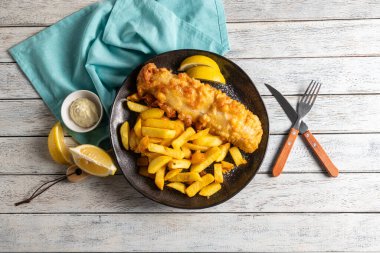 Tabakta, taze limon dilimleri ve küçük bir kase kremalı sosla süslenmiş altın kızartmaların yanında bol miktarda balık fileto servis ediliyor. Kırsal ahşap masa sıcaklık katıyor..