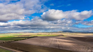 Uçsuz bucaksız tarım arazileri dramatik bir gökyüzünün altında uzanır. Tüylü bulutlar taze toprak üzerinde gölgeler yaratır. Sahne, doğal tuvalin dingin güzelliğini yakalar..
