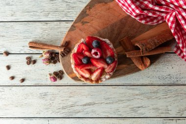 A vibrant fruit tart topped with strawberries, blueberries, and raspberries sits on a rustic wooden board. Coffee beans and cinnamon sticks lie nearby, enhancing the inviting atmosphere.