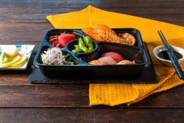 Colorful sushi and sashimi presented on a black tray, accompanied by fresh edamame and garnishes. The warm ambiance is enhanced by a bright yellow napkin and natural wood textures.