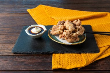 A plate of golden, crispy fried chicken sits on a rustic slate platter, accompanied by a small bowl of savory dipping sauce and a dollop of creamy condiment, all on a vibrant yellow cloth.
