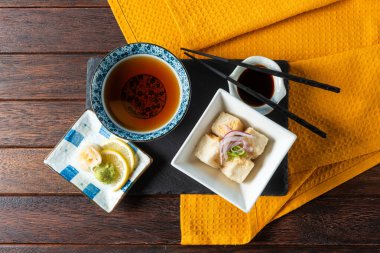A beautifully arranged dining setup featuring a bowl of tofu delicately garnished, accompanied by a steaming cup of tea and colorful condiments, all placed on a vibrant yellow tablecloth.