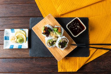 Delicious sushi rolls are neatly arranged on a wooden board, accompanied by a small bowl of dark sauce and garnished with wasabi and fresh lemon slices.