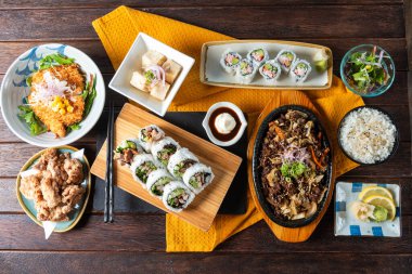 A vibrant spread featuring assorted sushi rolls, crispy tempura, and stir-fried delicacies set on a warm wooden table. A feast perfect for food lovers enjoying a shared meal.