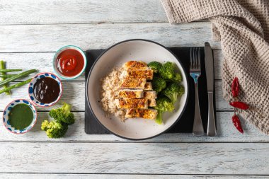 Colorful dish features grilled chicken strips resting on a bed of rice, accompanied by vibrant broccoli and an array of dipping sauces, all set on a rustic wooden table.