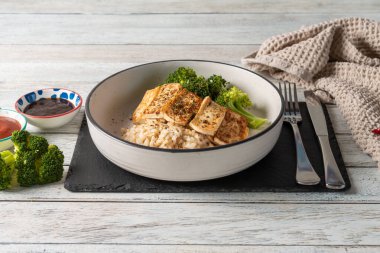 A vibrant bowl of fluffy rice topped with seared tofu and fresh broccoli sits elegantly on a rustic table. Accompanying dipping sauces add bursts of flavor to this nutritious dish.