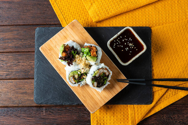 A beautifully arranged sushi platter featuring various rolls on a wooden board. A small bowl of dipping sauce accompanies the dish, all set against a vibrant orange napkin.