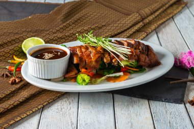Taze sebzeler ve küçük bir kase baharatlı sosla çevrili ızgara et parçaları beyaz bir tabakta dinleniyor. Bir dilim limon ve süsleme bu iştah açıcı yemeğe renk katar..
