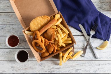 Canlı bir kızarmış yiyecek seçimi, içinde altın kızartmalar, soğan halkaları ve çıtır balık ve tavuk parçaları bulunan bir karton yemek kutusunu doldurur..