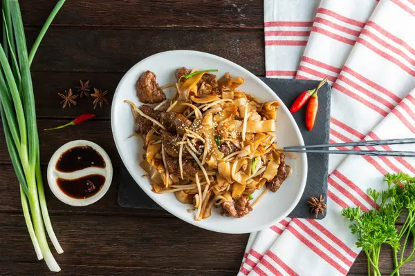 Lezzetli sığır eti, taze sebzeler ve aromatik baharatlarla dolu canlı bir tabak koyu ahşap bir masada oturur. Yemek çubukları sosun yanında dinlenir, aşçılık araştırmalarına davetiye çıkarır..