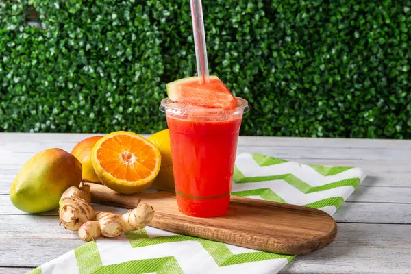 Renkli bir içecek taze meyve aromalı, parlak portakallar ve lezzetli mangoların arasında ahşap bir tahtanın üzerinde oturuyor. Yeşillik canlı atmosferi arttırır, güneşli bir gün için mükemmeldir..