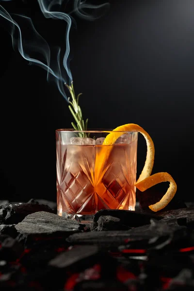 Old-fashioned cocktail with ice, orange peel, and rosemary. Whiskey with a burning twig of rosemary on a dark background.