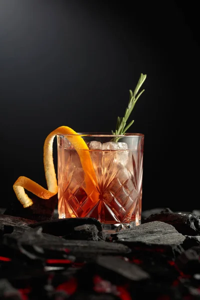 Old-fashioned cocktail with ice, orange peel, and rosemary. Glass with a cocktail on burning charcoal. Copy space.