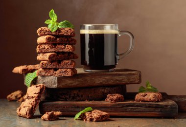 Pieces of fresh brownie with mint and a cup of black coffee on a rustic brown background.