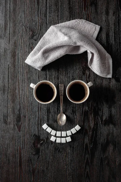 Coffee concept on a black wooden background. Coffee cups, towel, and sugar. The composition is like a human face. Copy space.