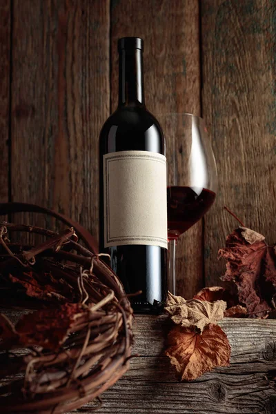Bottle and glass of red wine on an old wooden table with dried-up vine leaves. On a bottle old empty label. Focus on a label.
