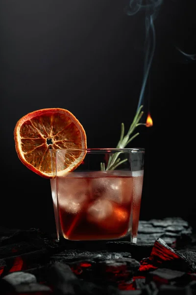 Old-fashioned cocktail with ice, dried orange slice, and rosemary. Whiskey with rosemary and beautiful swirls of smoke on a black background.