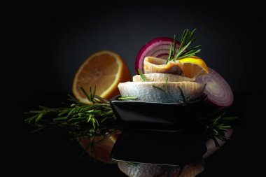 Limonlu, kırmızı soğanlı ve biberiyeli ringa fileto. Siyah yansıtıcı arka planda ringa balığı olan siyah bir tabak. Boşluğu kopyala.