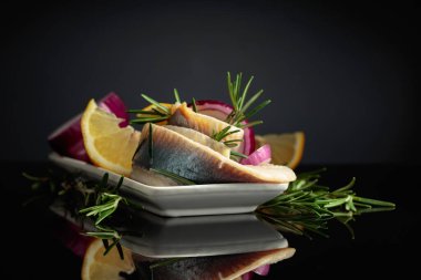 Limonlu, kırmızı soğanlı ve biberiyeli ringa fileto. Siyah yansıtıcı arka planda ringa balıklı beyaz tabak..