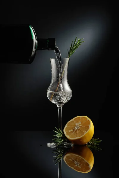 アンティークの瓶から小さなガラスにジンを注ぐ 黒の反射的な背景にレモンとローズマリーで飲む — ストック写真