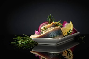 Limonlu, kırmızı soğanlı ve biberiyeli ringa fileto. Siyah yansıtıcı arka planda ringa balıklı beyaz tabak..