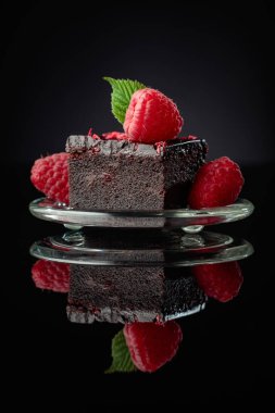 Chocolate cake garnished with fresh raspberries on a black reflective background.