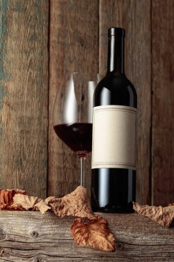 Kurumuş asma yaprakları olan eski ahşap bir masada şarap şişesi ve bir bardak kırmızı şarap. Eski boş bir şişede. Bir etikete odaklan.