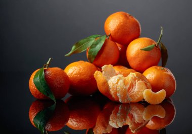 Siyah yansıtıcı arka planda yeşil yapraklı mandalinalar veya mandalinalar.