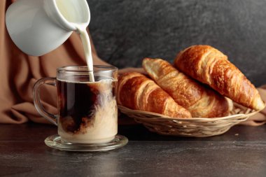 Freshly baked croissants and coffee with cream. Pouring creme in a glass cup of coffee.