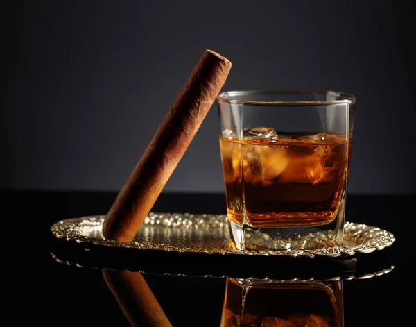 Whiskey with ice and cigar on a black reflective background.