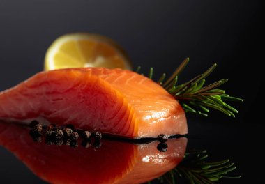 Smocked salmon piece with rosemary, lemon, and peppercorn on a black reflective background.