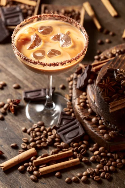 Irish coffee and cream cocktail in a glass with ice. Coffee beans, cinnamon, anise, and pieces of chocolate are scattered on the table.