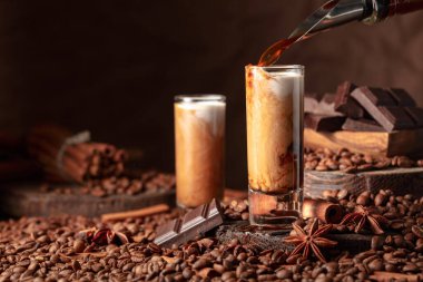 Irish cream and coffee liquor is poured from a bottle into a glass. Coffee beans, cinnamon, anise, and pieces of bitter chocolate are scattered on the table.