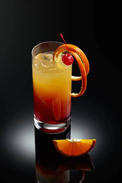 Cocktail tequila sunrise with a cherry and orange slice on a black reflective background.