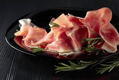 Italian prosciutto or Spanish jamon with rosemary on a black plate.