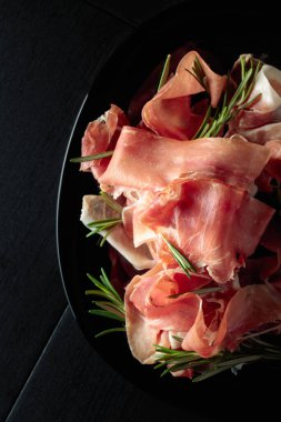 Italian prosciutto or Spanish jamon with rosemary on a black plate. Top view.