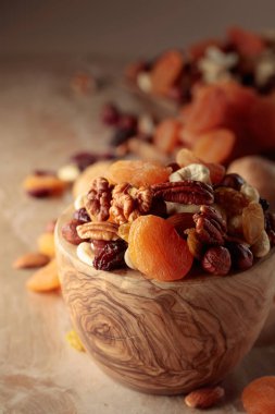 Dried fruits and nuts on a beige ceramic table. The mix of nuts, apricots, and raisins in a wooden bowl.