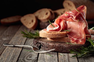 Prosciutto with bread and rosemary on an old wooden table. Copy space.