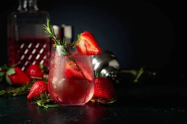 Cocktail gin tonic with ice, strawberries, and rosemary.  An iced refreshing drink in misted glass on a dark background. Copy space.