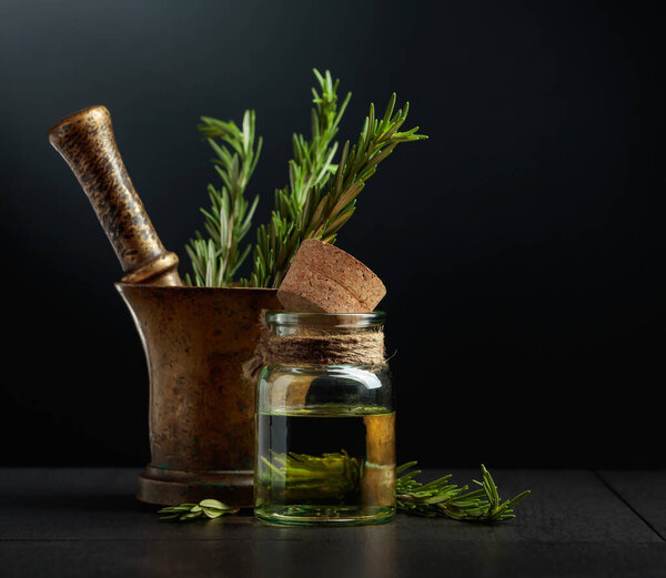 Bottle of rosemary aromatherapy oil extract with fresh rosemary branches and old brass mortar. Black background with copy space.