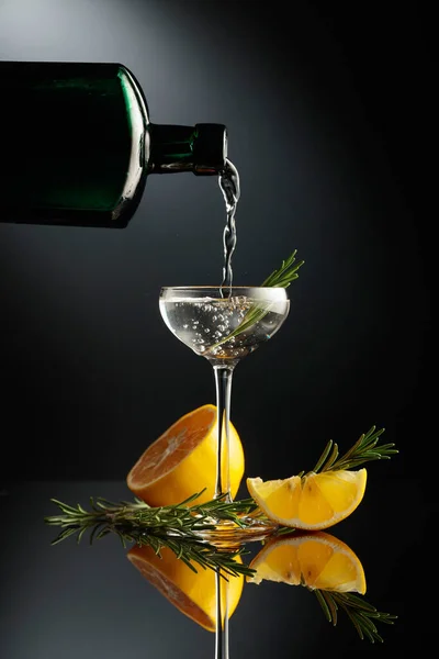 黒ガラスのアンティークボトルから小さなガラスにジンを注ぐ 黒の反射的背景にレモンとローズマリーとアルコール飲料 — ストック写真