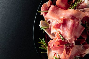 Italian prosciutto or Spanish jamon with rosemary on a black plate. Top view.
