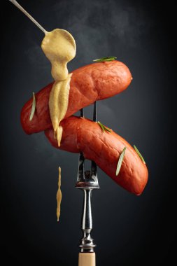 Boiled sausages with rosemary and mustard on a fork. Hot sausages with smoke on a black background.