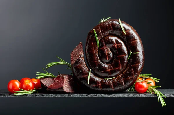 Traditional European black pudding or blood sausage with rosemary and tomatoes on a black background. Copy space.
