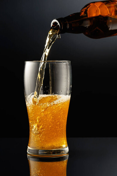 Pouring beer from a bottle into a glass. Glass of beer on a black reflective background.
