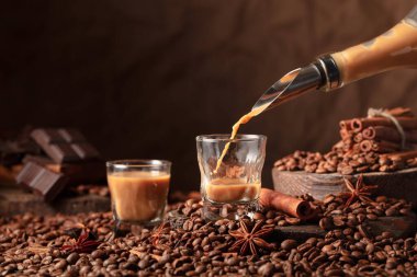 Irish cream and coffee liquor is poured from a bottle into a glass. Coffee beans, cinnamon, anise, and pieces of bitter chocolate are scattered on the table.