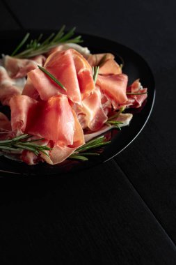 Italian prosciutto or Spanish jamon with rosemary on a black plate.