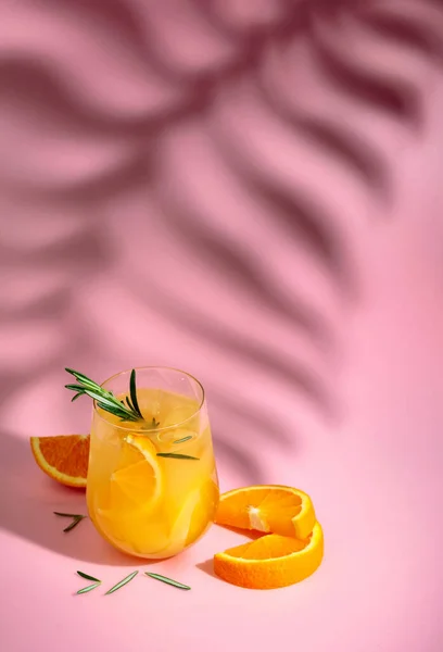 stock image Summer cocktail with orange, rosemary, and ice. Drink on pink background with palm leaf shadow.