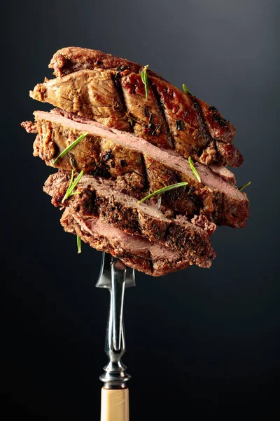 黒い背景にローズマリーとライビービーフステーキを焼きます ローズマリーを振りかけたフォークの上の牛ステーキ — ストック写真